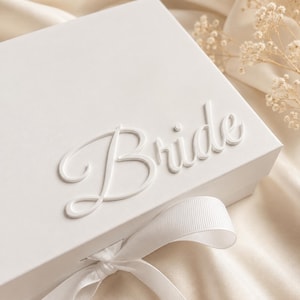 Könnte beinhalten: Eine rechteckige weiße Geschenkbox mit dem Wort Bride in erhabener weißer Schreibschrift auf dem Deckel. Die Box ist mit einem weißen Ripsband verschlossen, das vorne zu einer Schleife gebunden ist, was für eine klare und monochrome Optik sorgt.