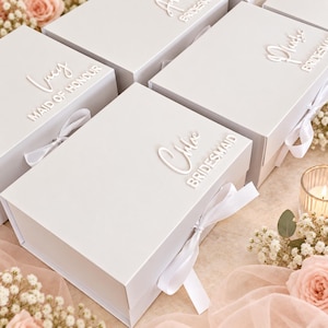 May include: A collection of white rectangular gift boxes with magnetic closures and white ribbon ties. Each box features raised white lettering displaying names such as Lucy, Chloe, and Phoebe, along with titles like Maid of Honour and Bridesmaid.