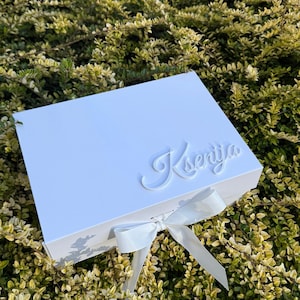 Puede incluir: Una caja de regalo rectangular de color blanco con un lazo blanco atado en la parte frontal. La tapa est&aacute; decorada con el nombre Ksenija en una fuente cursiva blanca en relieve. La caja tiene un acabado liso y un dise&ntilde;o estructurado.
