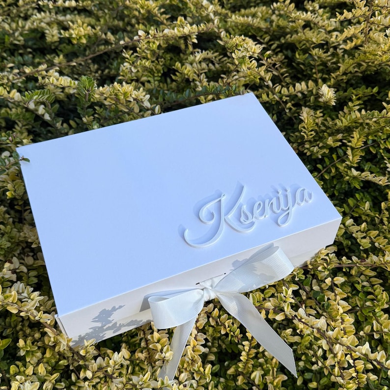 Puede incluir: Una caja de regalo rectangular de color blanco con un lazo blanco atado en la parte frontal. La tapa est&aacute; decorada con el nombre Ksenija en una fuente cursiva blanca en relieve. La caja tiene un acabado liso y un dise&ntilde;o estructurado.