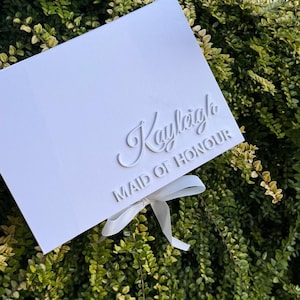 Puede incluir: Una caja de regalo rectangular blanca con el nombre Kayleigh en una fuente cursiva en relieve y el texto MAID OF HONOUR en una fuente sans-serif en relieve. Una cinta blanca est&aacute; atada en un lazo en la parte inferior central de la tapa.