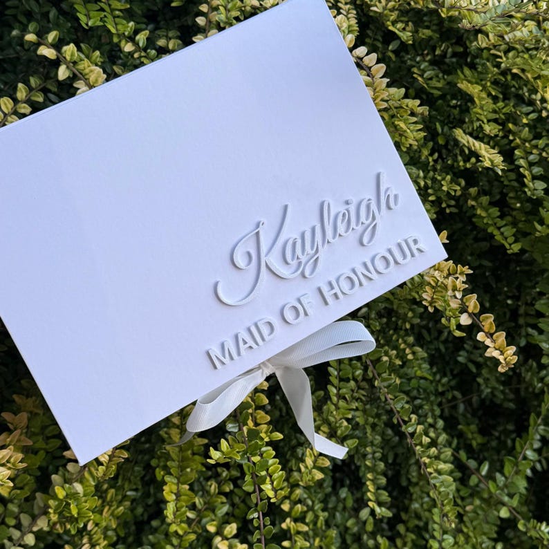 Puede incluir: Una caja de regalo rectangular blanca con el nombre Kayleigh en una fuente cursiva en relieve y el texto MAID OF HONOUR en una fuente sans-serif en relieve. Una cinta blanca est&aacute; atada en un lazo en la parte inferior central de la tapa.