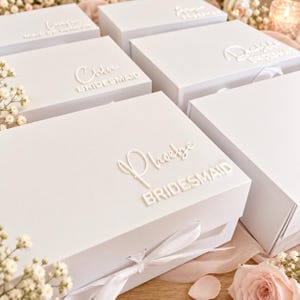 May include: A collection of white rectangular gift boxes with white ribbon closures. Each box features raised white lettering on the lid, displaying names such as Phoebe, Chloe, and Deirdre, along with titles like Bridesmaid and Maid of Honour.