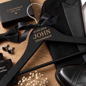 Pode incluir: Um cabide de madeira preta com um gancho de metal prateado e um laço de fita de cetim preto. O cabide está gravado com o texto dourado 03.09.2026, JOHN e GROOMSMAN. É exibido ao lado de um paletó preto, uma gravata borboleta e abotoaduras.
