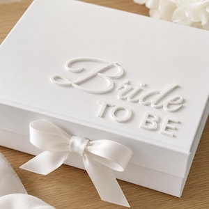 Könnte beinhalten: Eine rechteckige weiße Geschenkbox mit dem erhabenen weißen Schriftzug "Bride to be" auf dem Deckel. Die Box ist mit einem glatten weißen Satinband verschlossen, das vorne mittig zu einer Schleife gebunden ist und ein schlichtes, einfarbiges Design.