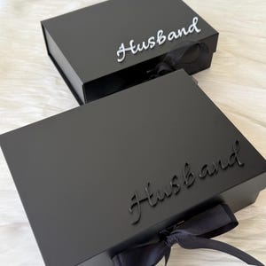 May include: Two rectangular black gift boxes with hinged lids and black ribbon closures. The top box features the word Husband in white script lettering, while the bottom box displays the word Husband in black script lettering on the lid.