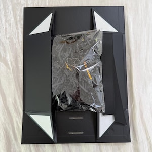 May include: A black rectangular gift box with magnetic closure flaps and white triangular accents at the corners. Inside, a clear plastic bag contains black shredded paper filler and a dark item with gold lettering visible through the plastic.