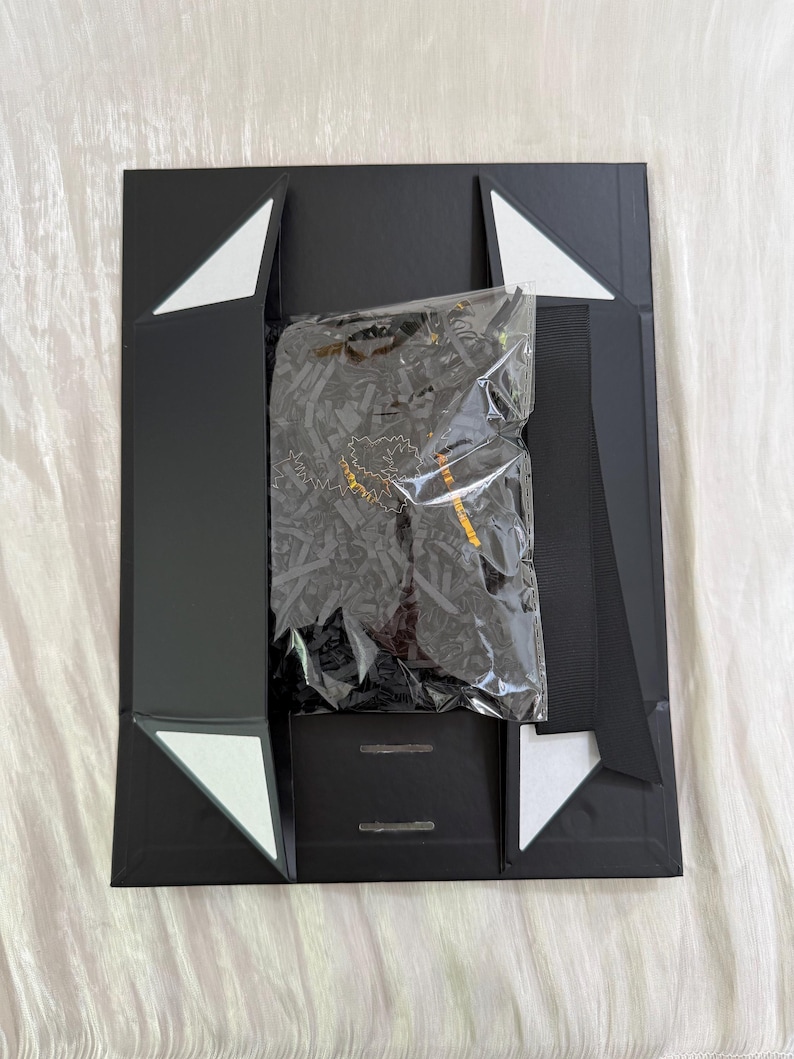 May include: A black rectangular gift box with magnetic closure flaps and white triangular accents at the corners. Inside, a clear plastic bag contains black shredded paper filler and a dark item with gold lettering visible through the plastic.