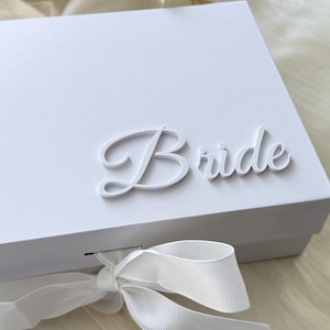 May include: A white rectangular gift box featuring the word Bride in raised white script lettering on the lid. The box is secured with a white grosgrain ribbon tied in a bow at the front, providing a clean and simple presentation for the item.