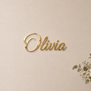 May include: A gold-coloured wall sign featuring the name Olivia in a cursive script font. The letters have a reflective, metallic finish and are mounted on a plain, light-coloured wall surface. The sign is positioned to the left of a flower arrangement.