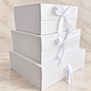 May include: A stack of three rectangular white gift boxes of varying sizes. Each box features a hinged lid and a white satin ribbon closure. The top two boxes are tied with a bow, while the bottom box has a single ribbon hanging down.