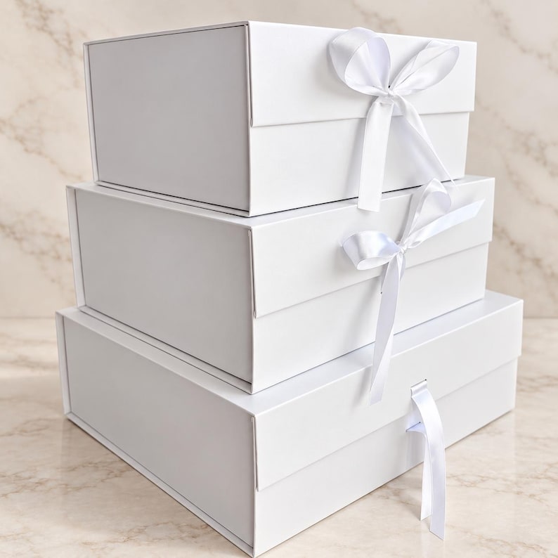 May include: A stack of three rectangular white gift boxes of varying sizes. Each box features a hinged lid and a white satin ribbon closure. The top two boxes are tied with a bow, while the bottom box has a single ribbon hanging down.