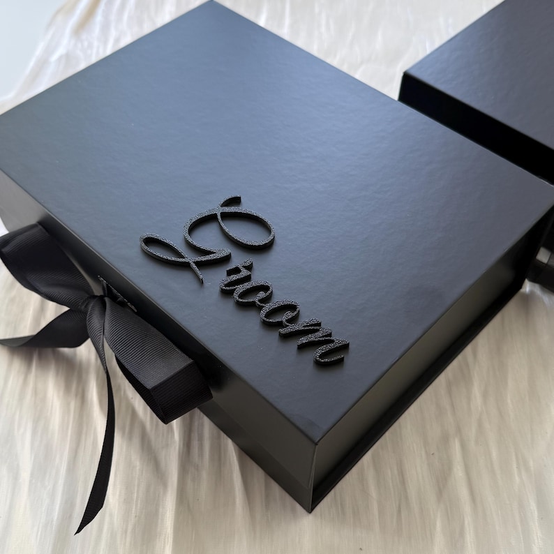 May include: A rectangular black gift box featuring a matte finish and a black ribbon closure on the left side. The top of the box is decorated with the word Groom in a raised, black, cursive script font. A second black box is visible in the corner.