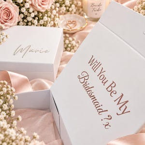 May include: A white gift box with the text Will You Be My Bridesmaid? x printed in rose gold, accompanied by a smaller white box featuring the name Marie in silver script, a candle labeled Bride Tribe, and pink roses with baby's breath flowers.