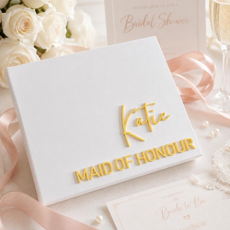 May include: A square white gift box featuring the name Katie in gold script lettering and the words MAID OF HONOUR in gold block lettering. The box is presented with soft pink ribbons, white roses, and printed bridal shower stationery.