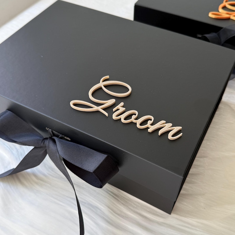 May include: A rectangular black gift box featuring a matte finish and a black ribbon tied in a bow on the front. The word Groom is written in a light beige, cursive, three-dimensional script font attached to the center of the box lid.