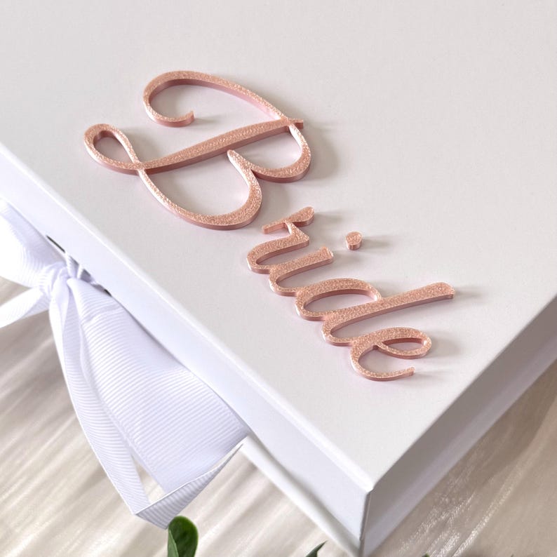 Puede incluir: Una caja de regalo rectangular blanca con una cinta blanca atada en un lazo en el lateral. La tapa est&aacute; decorada con la palabra Bride en una fuente cursiva tridimensional de color oro rosa brillante, fijada sobre la superficie superior.
