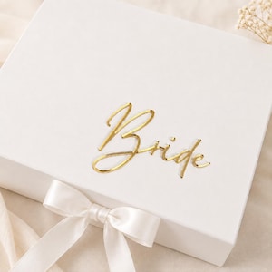 Op de afbeelding: Een vierkante witte geschenkdoos met de tekst Bride in goudkleurige metallic schrijfletters op het deksel. De doos is afgesloten met een wit satijnen lint dat aan de voorzijde tot een strik is gebonden voor een strakke en nette uitstraling.