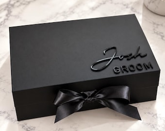 May include: A rectangular matte black gift box featuring a black satin ribbon tied in a bow at the front. The lid is decorated with the name Josh in a black script font and the word GROOM in black block lettering positioned below it.