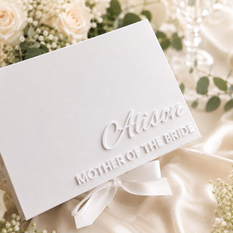 May include: A rectangular white gift box featuring the name Alison in raised script lettering and the text MOTHER OF THE BRIDE in raised block lettering. The box is finished with a white satin ribbon tied into a bow at the bottom center.