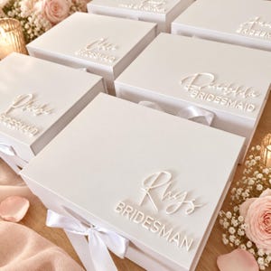 May include: A collection of white rectangular gift boxes, each featuring a white satin ribbon closure and raised white lettering on the lid. The text on the boxes includes individual names in script font followed by the word BRIDESMAID in block letters.