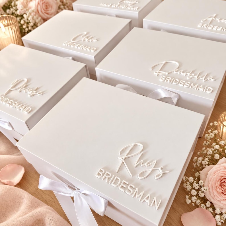 May include: A collection of white rectangular gift boxes, each featuring a white satin ribbon closure and raised white lettering on the lid. The text on the boxes includes individual names in script font followed by the word BRIDESMAID in block letters.