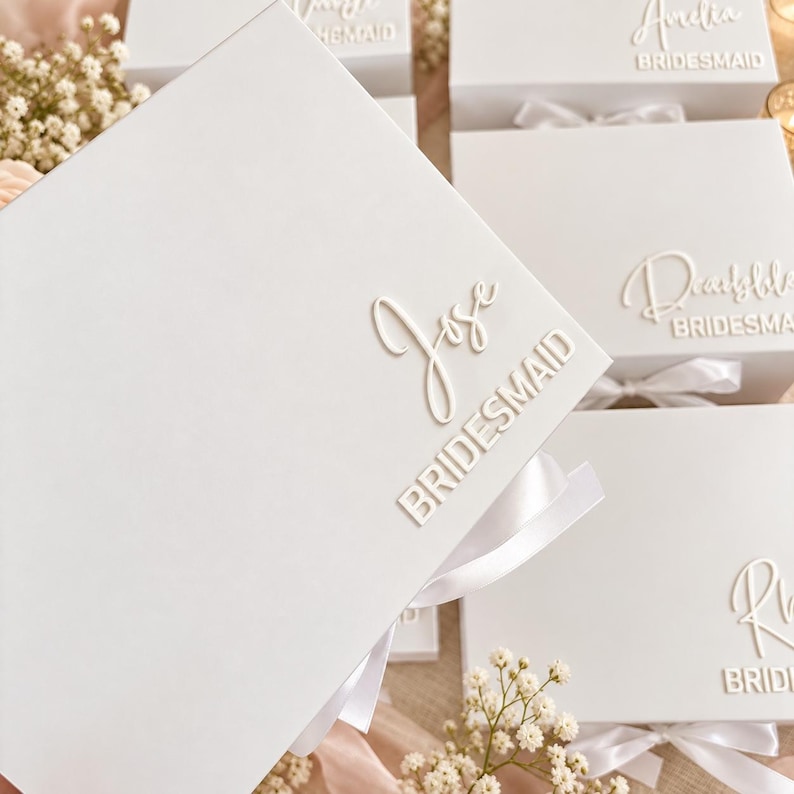 May include: A white rectangular gift box featuring a white satin ribbon and raised white lettering that reads Jose and BRIDESMAID. The box is one of several identical white boxes, each personalized with a name and the title BRIDESMAID.