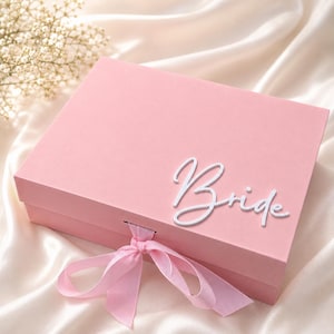 May include: A rectangular light pink gift box featuring the word Bride in white script lettering on the lid. The box is secured with a matching light pink ribbon tied in a bow at the front center, providing a clean and simple presentation.