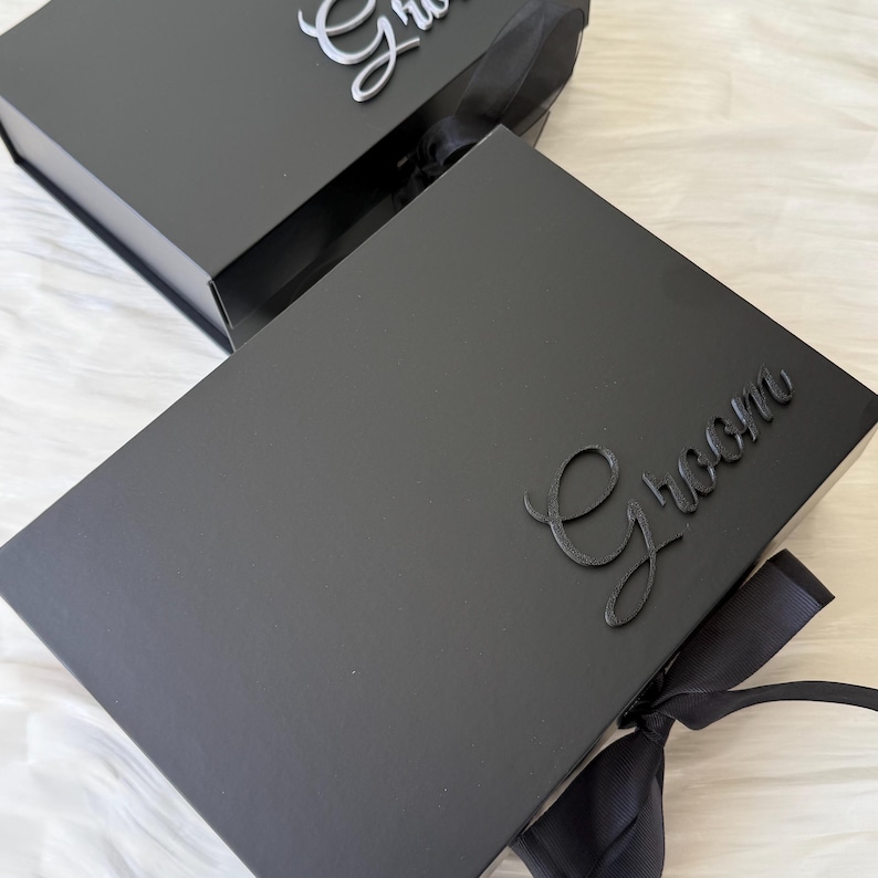 May include: Two rectangular black gift boxes with magnetic closures and black ribbon ties. The top box features the word Groom in raised black script lettering, while the partially visible box behind it displays a silver script word.