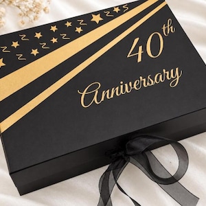 May include: A rectangular black gift box featuring gold foil accents, including diagonal stripes and a star pattern. The text 40th Anniversary is printed in gold script on the lid. A black ribbon is tied in a bow at the front edge of the box.