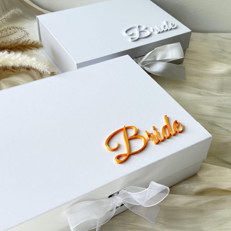 Puede incluir: Dos cajas de regalo rectangulares blancas con cierres magn&eacute;ticos y lazos de cinta blanca transparente. La caja frontal presenta la palabra Bride en letras cursivas naranjas en relieve, mientras que la trasera muestra la palabra Bride en blanco.
