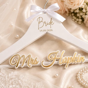 Op de afbeelding: Een witte houten kledinghanger met een witte satijnen strik aan de bovenkant. De hanger is bedrukt met de gouden tekst "Bride" en de datum "14.09.2026". Aan de onderste stang is een goudkleurig en wit naambordje met "Mrs. Hopkin" bevestigd.