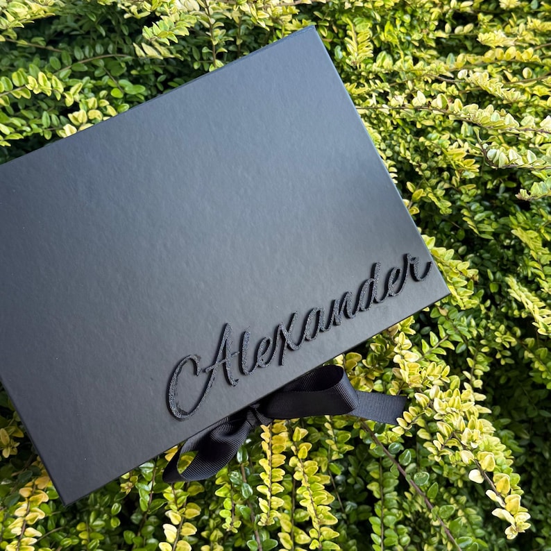May include: A rectangular black gift box featuring the name Alexander in raised black script lettering on the lid. A black ribbon is tied in a bow at the bottom edge of the box, providing a secure closure for the container.