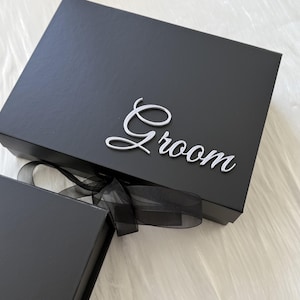 May include: A rectangular black gift box featuring the word Groom in a silver script font attached to the lid. The box is secured with a black sheer ribbon tied in a bow at the front, and a portion of a second black box is visible in the corner.