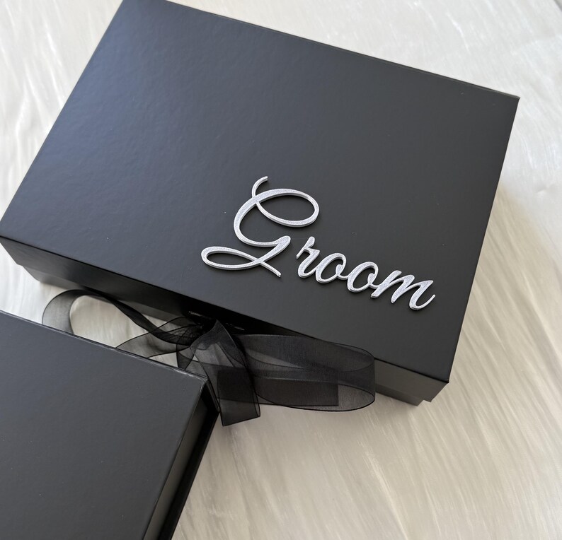 May include: A rectangular black gift box featuring the word Groom in a silver script font attached to the lid. The box is secured with a black sheer ribbon tied in a bow at the front, and a portion of a second black box is visible in the corner.