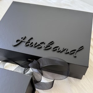 May include: A rectangular black gift box featuring the word Husband in a raised black script font on the lid. The box is tied with a sheer black ribbon that is tied into a bow at the front, and a smaller black box is visible in the foreground.