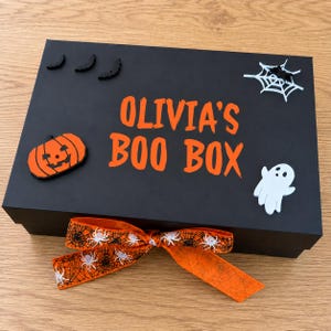 May include: A rectangular black gift box featuring the orange text OLIVIA'S BOO BOX. The lid is decorated with black bat silhouettes, a white spider web, an orange pumpkin, and a white ghost. An orange ribbon with a black spider web pattern is tied at the front.
