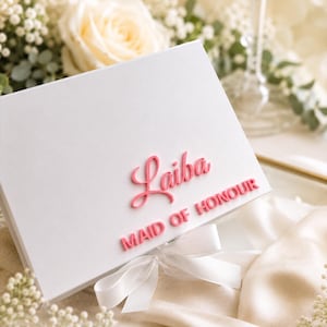 May include: A rectangular white gift box featuring the name Laiba in a pink cursive script and the text MAID OF HONOUR in pink block letters. The box is adorned with a white satin ribbon tied into a bow at the bottom center of the lid.