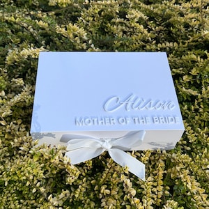 Puede incluir: Una caja de regalo rectangular blanca con un lazo de cinta blanca en la parte delantera. La tapa muestra el nombre Alison en una fuente cursiva en relieve, con el texto MOTHER OF THE BRIDE en una fuente sans-serif en relieve debajo del nombre.
