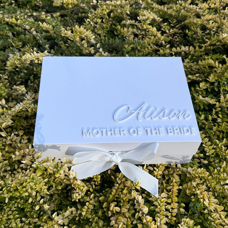 Puede incluir: Una caja de regalo rectangular blanca con un lazo de cinta blanca en la parte delantera. La tapa muestra el nombre Alison en una fuente cursiva en relieve, con el texto MOTHER OF THE BRIDE en una fuente sans-serif en relieve debajo del nombre.