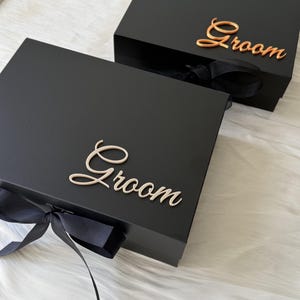May include: Two rectangular black gift boxes with lids, each featuring the word Groom in elegant script lettering. One box displays the text in a metallic rose gold finish, while the other features a light cream script. Both boxes have black ribbon bows.