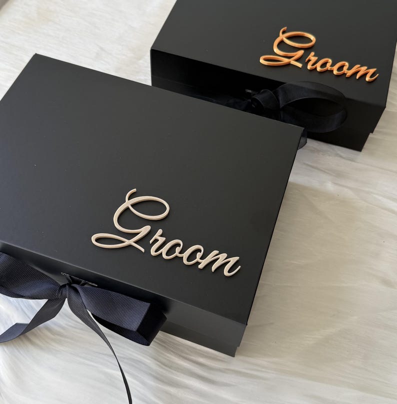 May include: Two rectangular black gift boxes with lids, each featuring the word Groom in elegant script lettering. One box displays the text in a metallic rose gold finish, while the other features a light cream script. Both boxes have black ribbon bows.