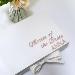 Puede incluir: Una caja de regalo rectangular blanca con el texto en oro rosa Mother of the Bride ANNA en la tapa. La caja est&aacute; cerrada con una cinta blanca atada en un lazo en el borde frontal. El dise&ntilde;o es limpio y sencillo con un acabado mate suave.