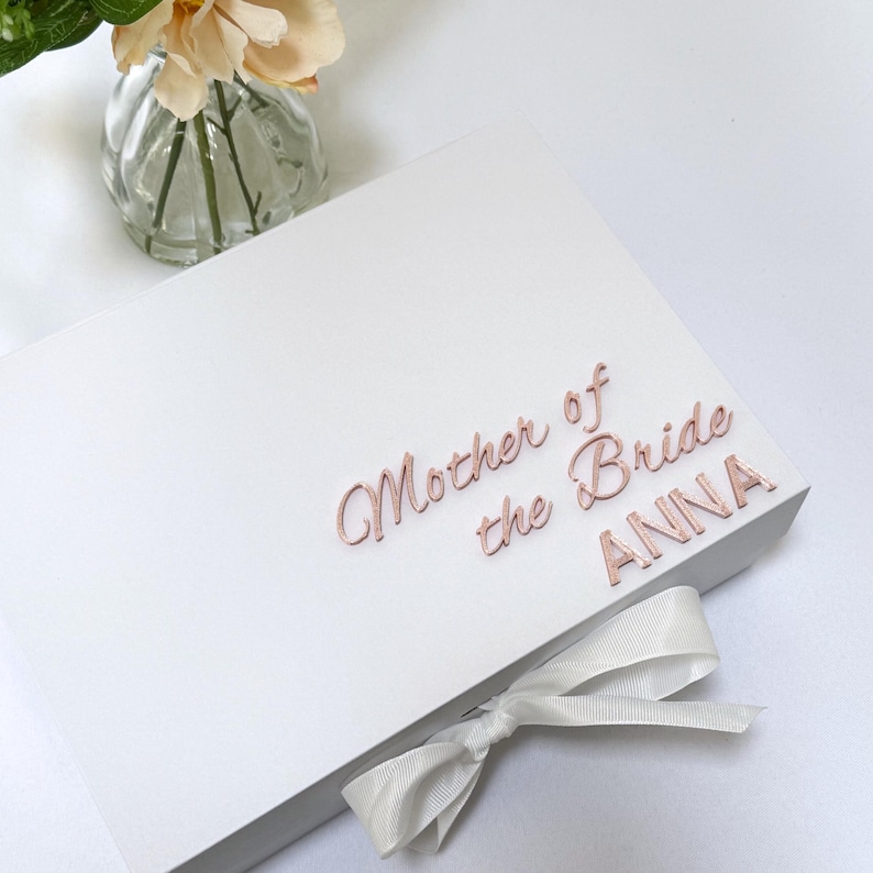 Puede incluir: Una caja de regalo rectangular blanca con el texto en oro rosa Mother of the Bride ANNA en la tapa. La caja est&aacute; cerrada con una cinta blanca atada en un lazo en el borde frontal. El dise&ntilde;o es limpio y sencillo con un acabado mate suave.
