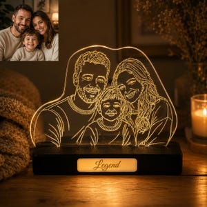 Op de afbeelding: Een gepersonaliseerd LED-nachtlampje met een gegraveerd lijnportret van een gezin van drie op een helder acrylpaneel. Het paneel staat op een rechthoekige zwarte voet met een verlicht voorpaneel waarop het woord Legend in scriptlettertype staat.