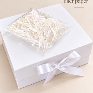 Puede incluir: Una caja de regalo cuadrada de color blanco con un lazo de sat&eacute;n blanco en la parte frontal. Una bolsa de pl&aacute;stico transparente con papel de relleno blanco triturado descansa sobre la caja. El texto "Comes with filler paper" aparece arriba.