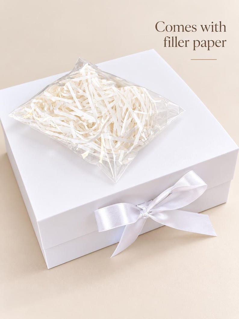 Puede incluir: Una caja de regalo cuadrada de color blanco con un lazo de sat&eacute;n blanco en la parte frontal. Una bolsa de pl&aacute;stico transparente con papel de relleno blanco triturado descansa sobre la caja. El texto "Comes with filler paper" aparece arriba.