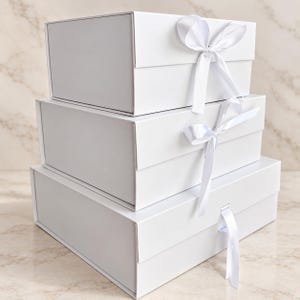 Puede incluir: Una pila de tres cajas de regalo rectangulares blancas de diferentes tama&ntilde;os. Cada caja cuenta con una tapa abatible y un cierre de cinta de sat&eacute;n blanca. Las dos cajas superiores tienen un lazo, mientras que la inferior tiene una cinta.