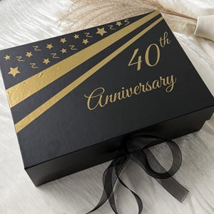 Könnte beinhalten: Eine rechteckige schwarze Geschenkbox mit Goldfolienakzenten, darunter diagonale Streifen und ein Muster aus kleinen Sternen. Der Text 40th Anniversary ist in goldener Schrift aufgedruckt. Ein schwarzes, transparentes Band ist vorne gebunden.