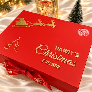 May include: A rectangular red gift box featuring gold foil accents including a silhouette of Santa in a sleigh with reindeer, a stylised Christmas tree, and the text "HARRY'S Christmas EVE BOX" with a "NORTH POLE FROM SANTA" circular seal.